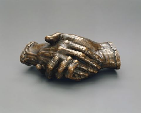 Sculpture. Bronze cast of two right hands holding each other. The hands cut off at the wrists, and one is edged in a straight cuff and the other in scalloped cuff. In this view, one rests along the palm of the other. Veins on the back of one hand and on the underside of the wrist of the other are visible. The patina is dark brown on most of the surface with a few brighter areas of gold.