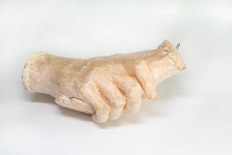 Sculpture. Plaster cast of two right hands holding each other. The hands cut off at the wrists, and one is edged in a straight cuff and the other in scalloped cuff. In this view, the fingers of one hand wrap around the outer edge of the other.